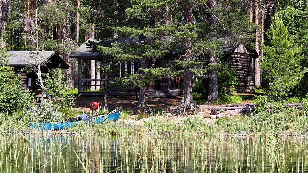 A log hut surrounded by pine forest. At front there is a hiker and a boat on the reedy shore.